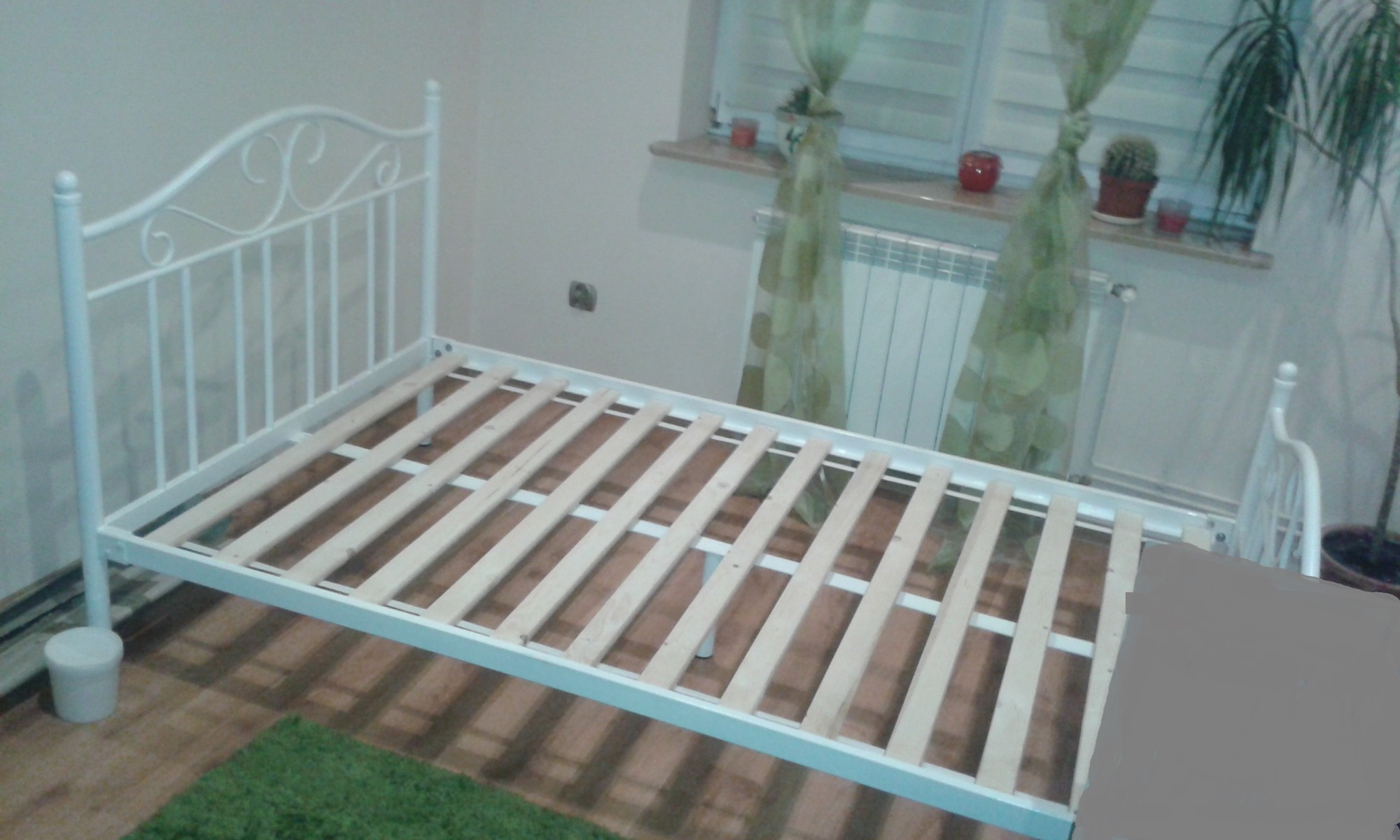 Białe metalowe łóżko z drewnianym stelażem, widoczne w pokoju z zielonym dywanem i zasłonami.