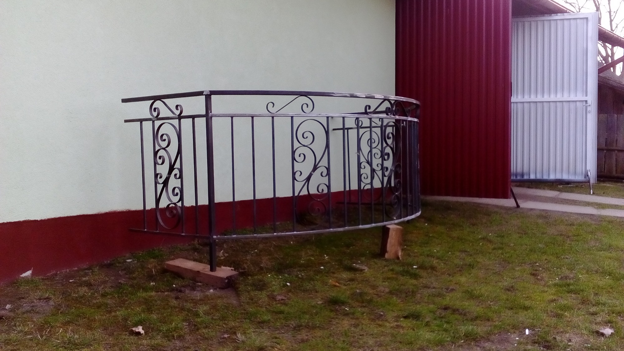 Kuta, czarna balustrada ochronna z ozdobnymi wzorami, zamontowana przy budynku z zielonkawą ścianą i czerwoną podmurówką, podparta drewnianymi klinami, z otwartą bramą garażową w tle.