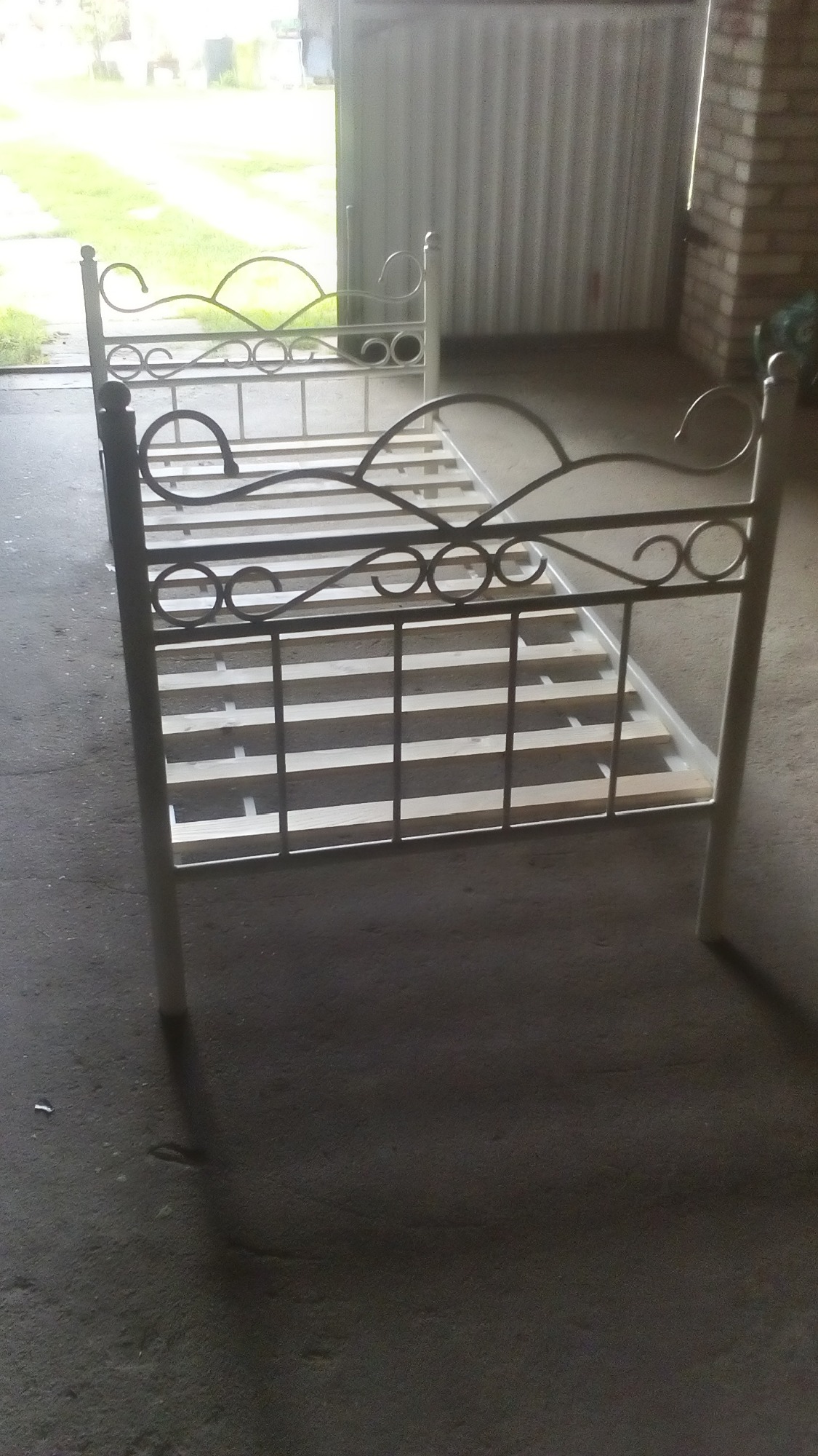 Metalowa konstrukcja łóżka z ozdobnymi, kręconymi elementami, pomalowana na biało, ustawiona w garażu z betonową podłogą.