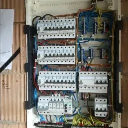 Otwarta skrzynka rozdzielcza z zainstalowanymi bezpiecznikami i okablowaniem, zamontowana na ścianie z cegły dziurawki.