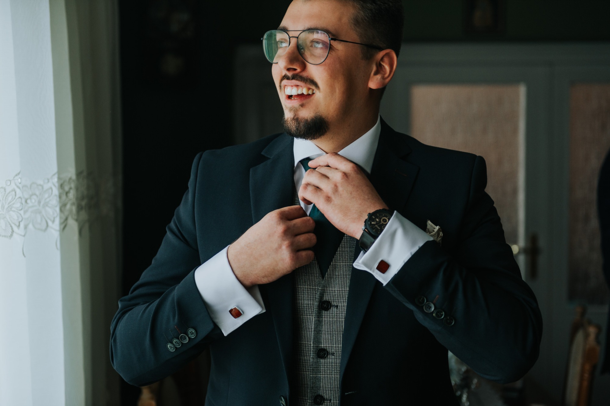 Uśmiechnięty mężczyzna w okularach, dopinający krawat do eleganckiego garnituru z kamizelką w kratę i mankietami z bordowymi spinkami, spogląda w bok przy jasnym oknie.