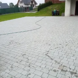 Umyta, szara kostka brukowa na podjeździe domu jednorodzinnego, z widocznym wężem ogrodowym i dwoma taczkami na trawniku w tle.