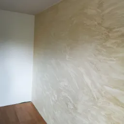 Ściana z efektem dekoracyjnym w odcieniach beżu i bieli, sąsiadująca z białą ścianą i brązową podłogą panelową.