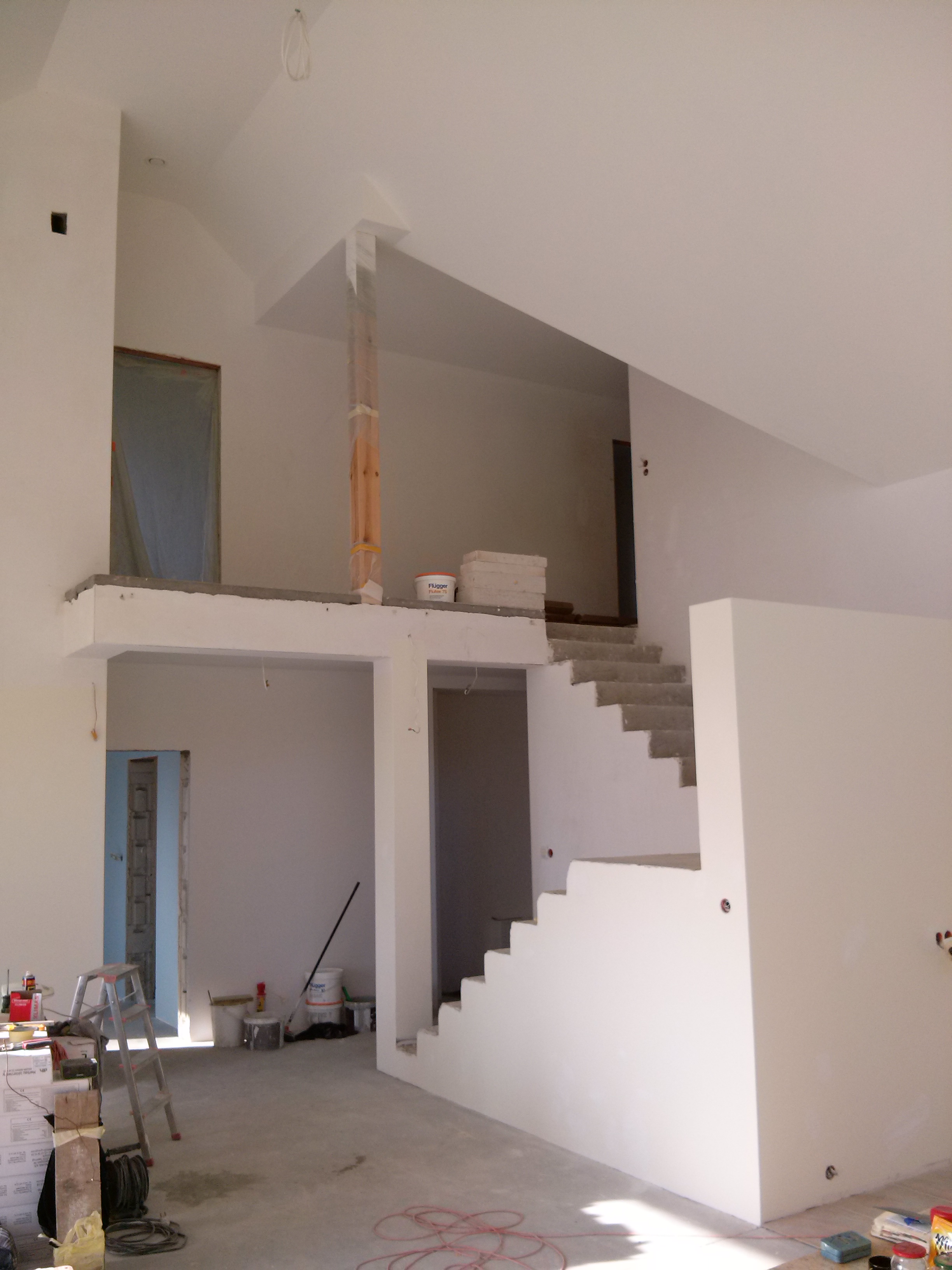 Wnętrze domu w trakcie remontu: białe, wysokie ściany, schody w stanie surowym, widoczne narzędzia i materiały budowlane, zabezpieczone drzwi folią.