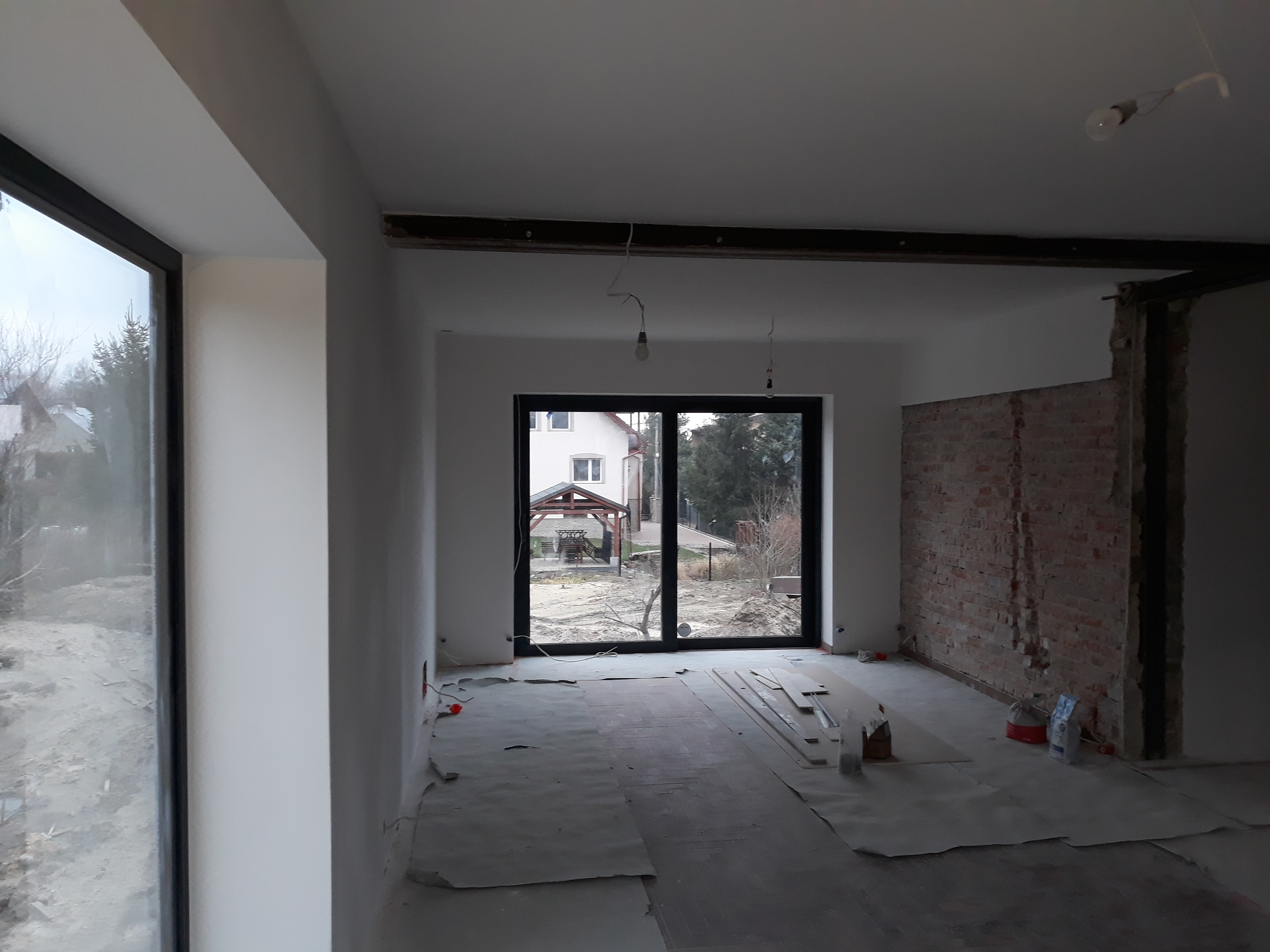 Wnętrze w trakcie remontu: świeżo pomalowane ściany na biało, odsłonięta cegła na fragmencie ściany, zabezpieczona podłoga, okna z czarnymi ramami, widoczne kable wystające ze ściany i żarówki...