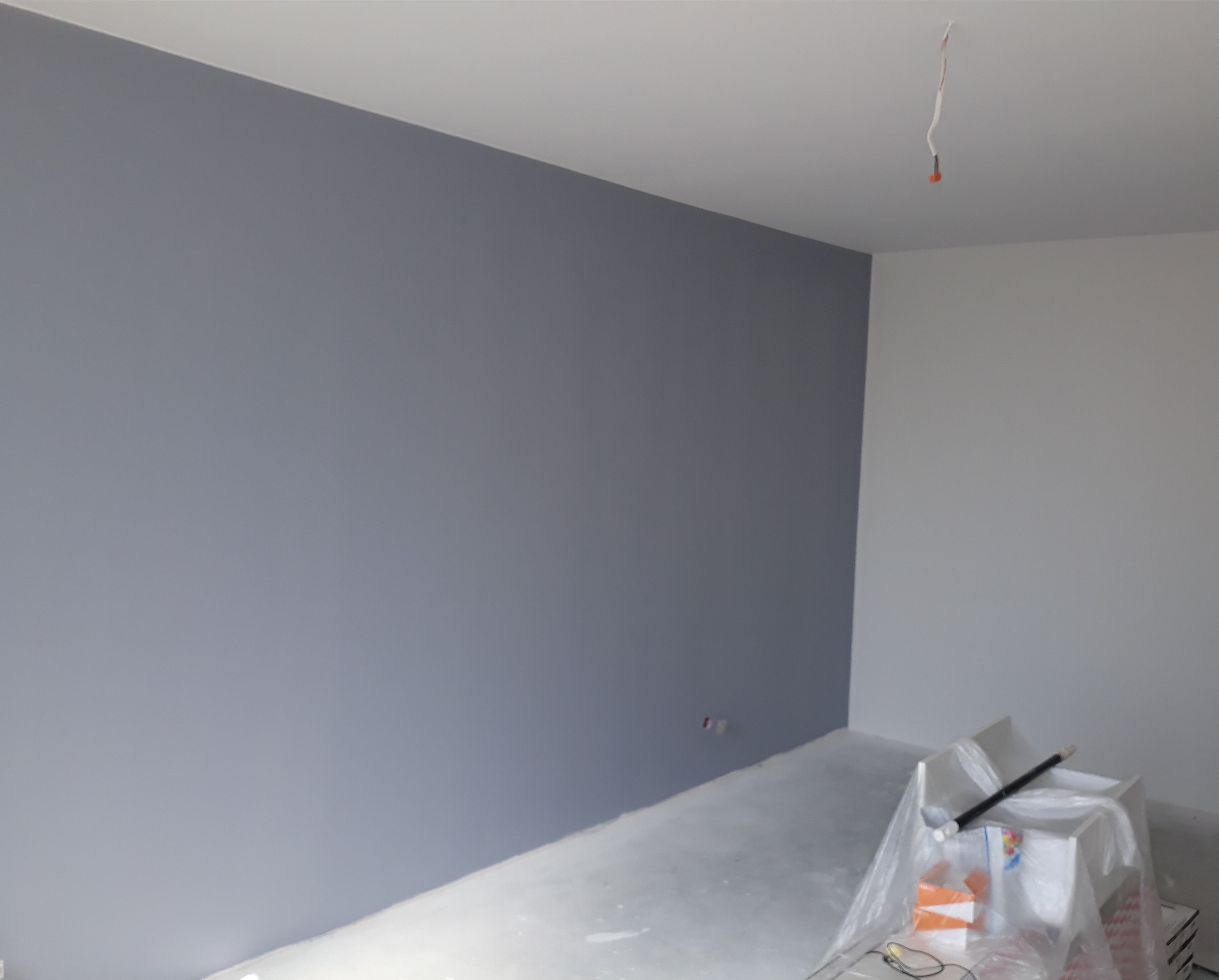 Świeżo pomalowane na szaro ściany w pokoju, z widocznymi kablami wystającymi z sufitu i zabezpieczonymi folią malarską akcesoriami w rogu pomieszczenia.