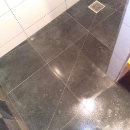 Krystian Napiorkowski - Szara, błyszcząca podłoga wyłożona dużymi płytkami w kabinie prysznicowej, widoczny odpływ liniowy i fragment buta osoby robiącej zdjęcie.