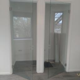 Krystian Napiorkowski - Dwa szklane skrzydła drzwiowe zamontowane w białych ościeżnicach, widoczne okucia i przezroczystość szkła, szara wykładzina podłogowa, jasne wnętrze z oknem w tle.