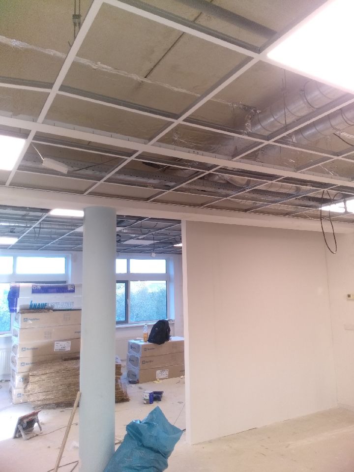 Wnętrze biura w trakcie remontu: widoczny sufit podwieszany z instalacją wentylacyjną, ściany przygotowane do malowania, materiały budowlane w kartonach i niebieski worek na podłodze.