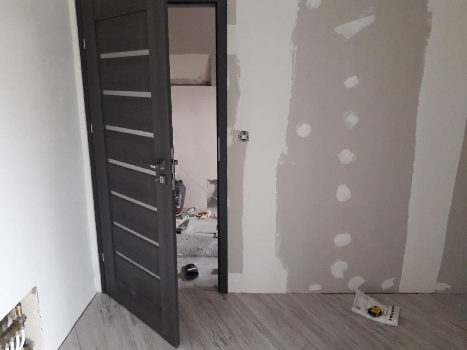 Wnętrze w trakcie remontu: widok na otwarte, grafitowe drzwi z ukośnymi, srebrnymi paskami, ściana z widocznymi łatami gipsowymi, podłoga imitująca drewno, w tle widoczny bałagan budowlany.
