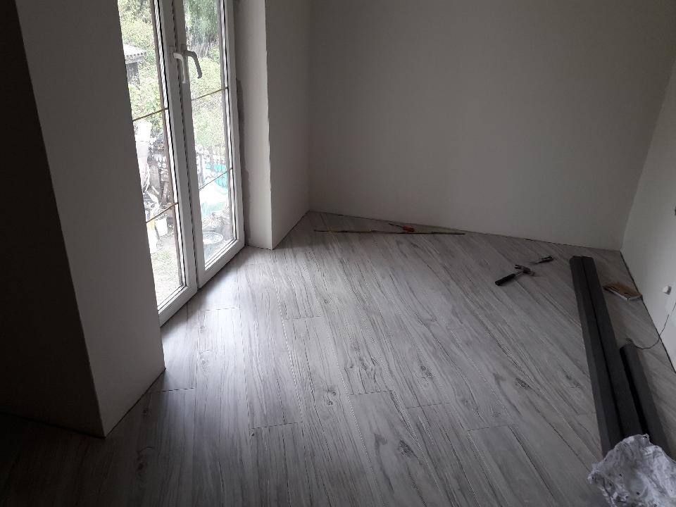 Pokój z nową podłogą z paneli imitujących drewno, widoczne narzędzia, listwy przypodłogowe i okno balkonowe z widokiem na zewnątrz.