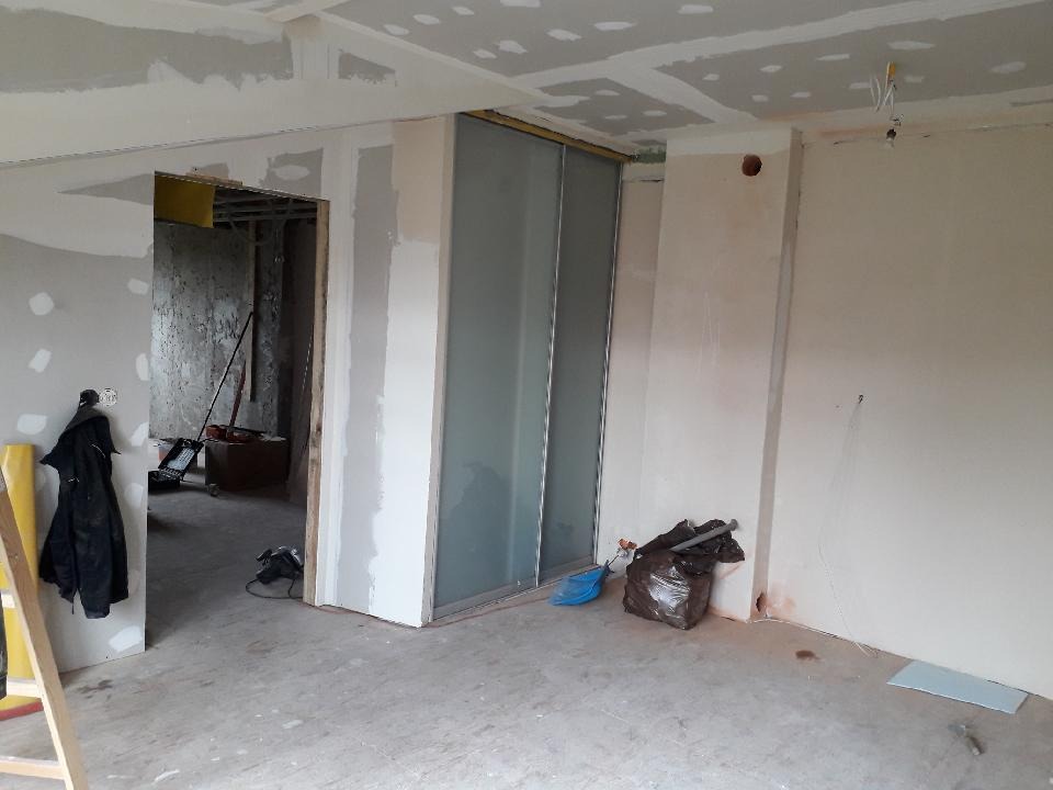 Wnętrze w trakcie remontu: ściany i sufit z płyt gipsowo-kartonowych, widoczne szpachlowanie i przygotowanie do malowania, przesuwne drzwi w metalowej ramie, narzędzia i worki z odpadami na podłodze.