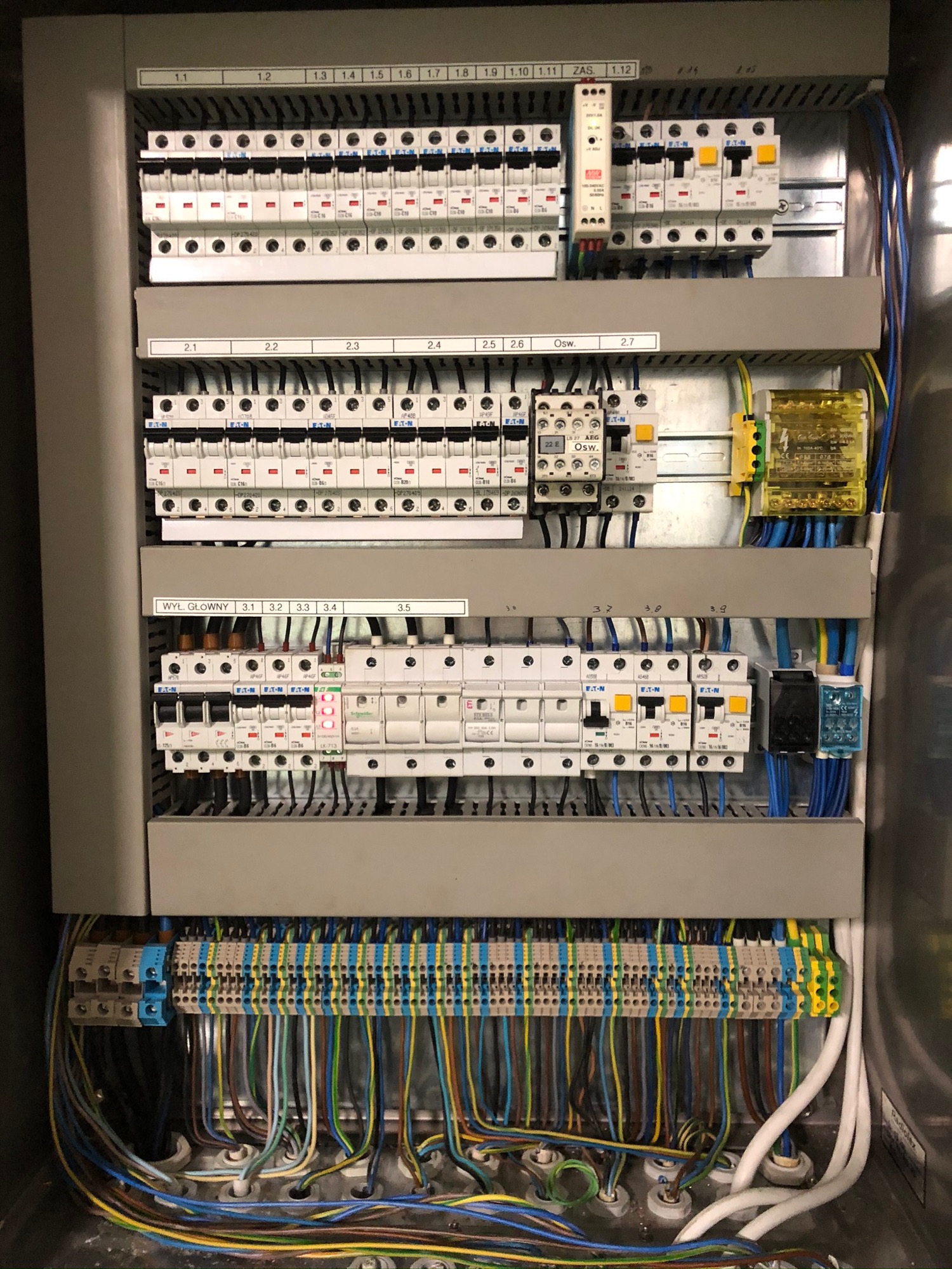 Rozdzielnica elektryczna z widocznymi bezpiecznikami, przewodami o różnych kolorach i oznaczeniami, wewnątrz metalowej obudowy, z naklejkami opisującymi poszczególne sekcje, oświetlona światłem...