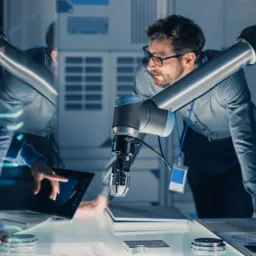 Dwóch inżynierów w laboratorium analizuje dane na tablecie, podczas gdy ramię robota precyzyjnie przetwarza metalowy element na podświetlonym stole.