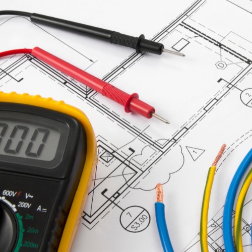 Multimetr z przewodami pomiarowymi leżący na planie instalacji elektrycznej, obok widoczne przewody w izolacji niebieskiej i żółto-zielonej.