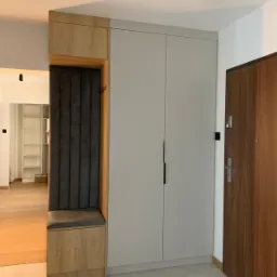 Nowoczesny przedpokój z zabudową: szafa w odcieniach szarości, drewniany panel z siedziskiem i wieszakami, oświetlenie punktowe na suficie. Minimalistyczny design.