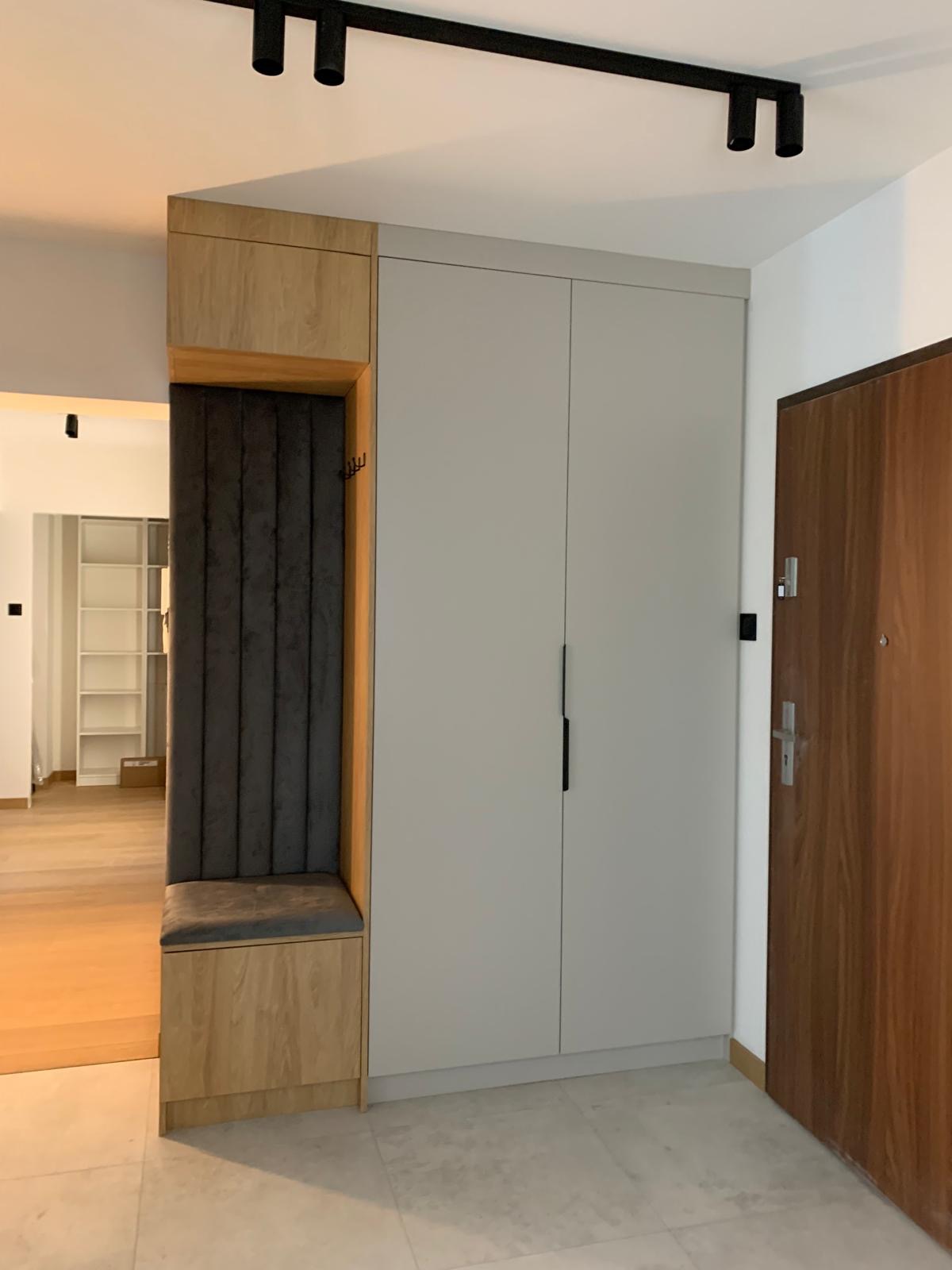 Nowoczesny przedpokój z zabudową: szafa w odcieniach szarości, drewniany panel z siedziskiem i wieszakami, oświetlenie punktowe na suficie. Minimalistyczny design.