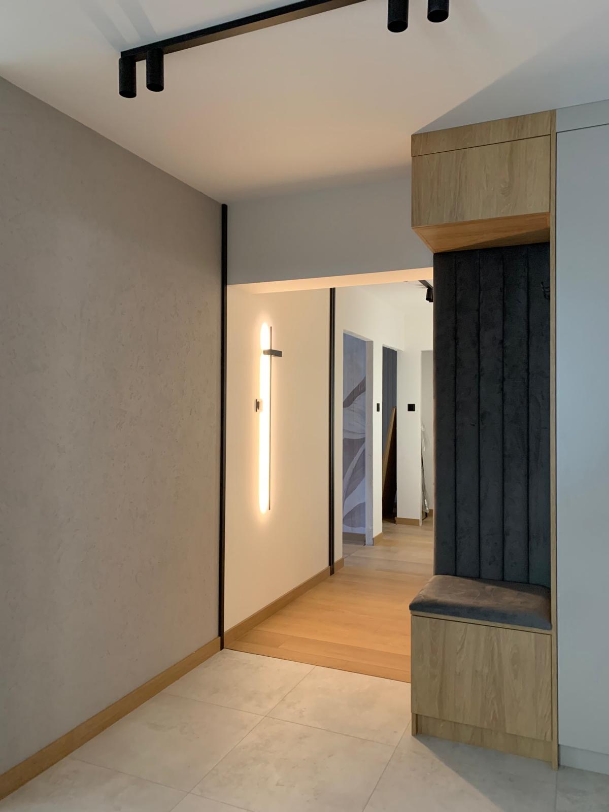 Nowoczesny hol z siedziskiem w zabudowie, oświetleniem LED i wykończeniem w odcieniach szarości i drewna. Minimalistyczny design.