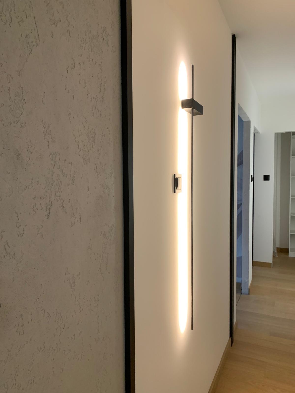 Nowoczesna lampa ścienna z pionową listwą LED i czarnym mocowaniem, na tle ściany z efektem dekoracyjnym i czarną ramą. Jasne, neutralne wnętrze.