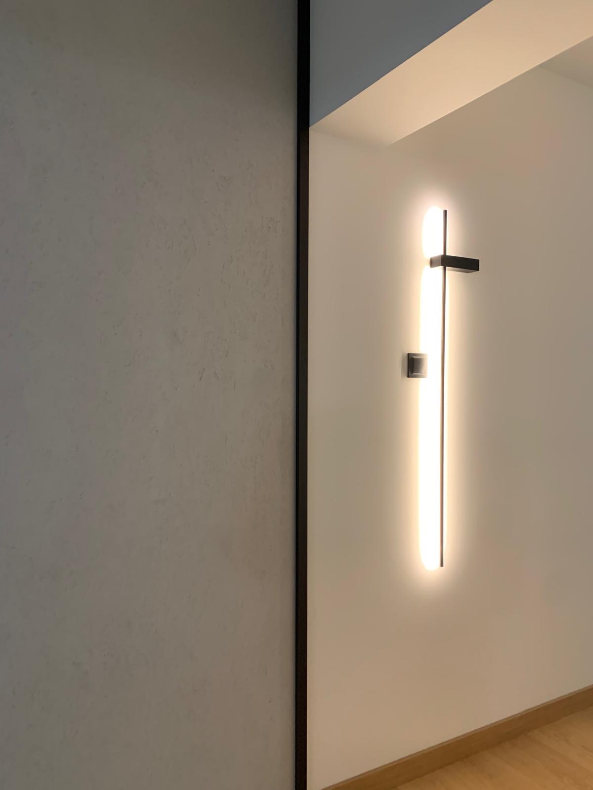 Minimalistyczna ściana z szarą, teksturowaną powierzchnią i pionową, podłużną lampą LED z czarnym akcentem, oświetlająca fragment parkietu.