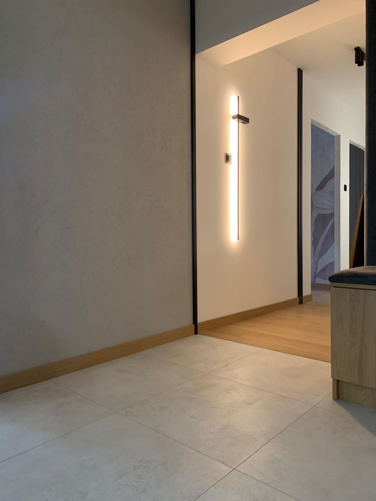 Minimalistyczne wnętrze: szara ściana z dekoracyjną fakturą, ledowa lampa pionowa i jasna podłoga z płytek, drewniane listwy przypodłogowe, widok na korytarz.
