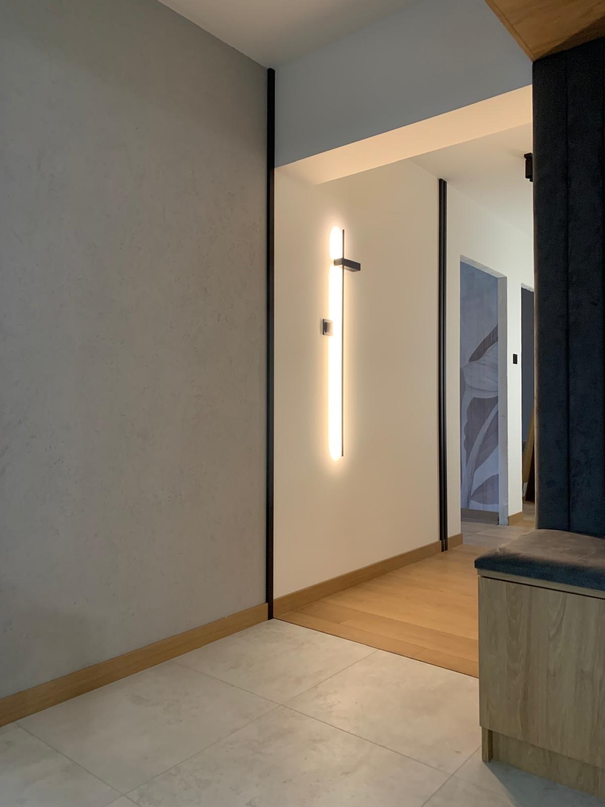 Nowoczesny przedpokój z szarą ścianą o fakturze, podświetlanym lustrem i drewnianą podłogą. Minimalistyczny design wnętrza z elementami wykończeniowymi.