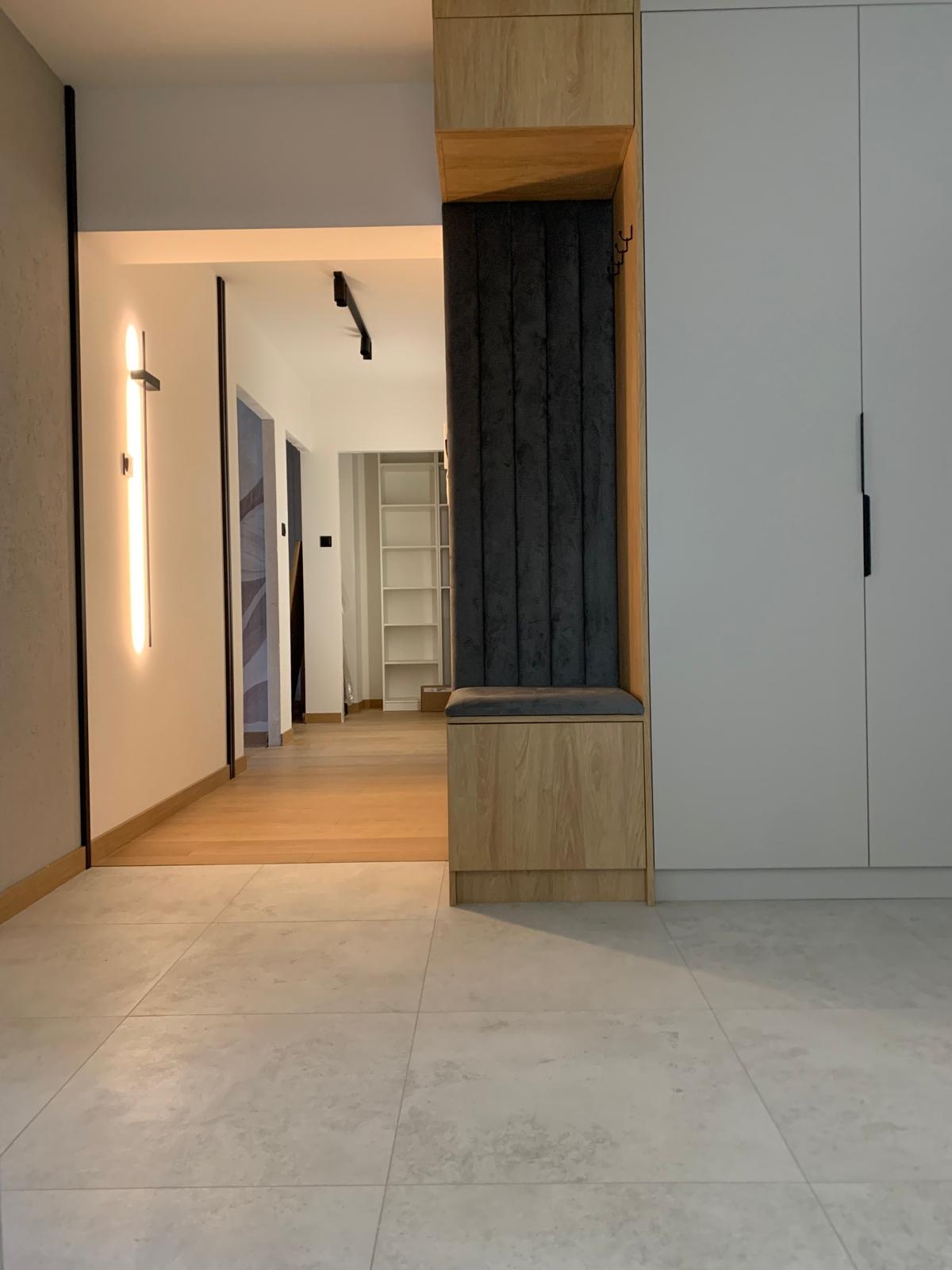 Nowoczesny przedpokój z zabudową meblową: siedzisko z tapicerowanym panelem, szafa w zabudowie, oświetlenie LED. Widok na korytarz z drewnianą podłogą i regałem.