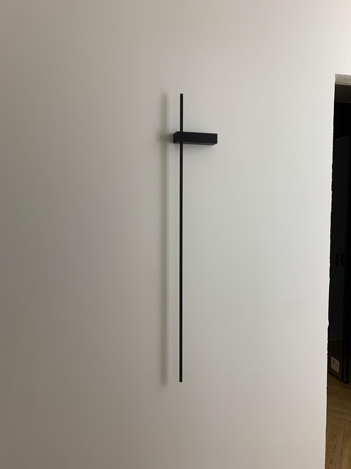 Minimalistyczna, czarna lampa ścienna w kształcie litery T, zamontowana na gładkiej, białej ścianie. Widoczny fragment drzwi w tle.