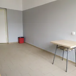 Szare ściany w pokoju po świeżym pomalowaniu, widoczna biała listwa przypodłogowa, brązowe płytki na podłodze i biurko na metalowych nogach z drewnianym blatem.