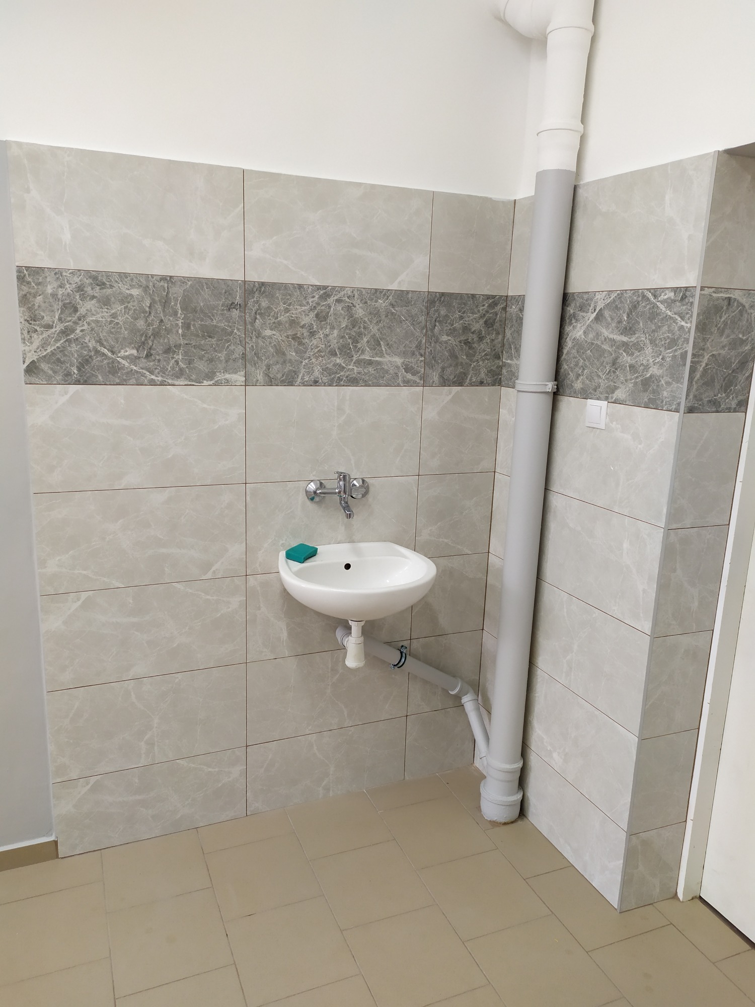 Mała umywalka z chromowaną baterią i zieloną gąbką na blacie, zamontowana na ścianie wyłożonej płytkami imitującymi marmur, z widoczną rurą kanalizacyjną i pionem wentylacyjnym.