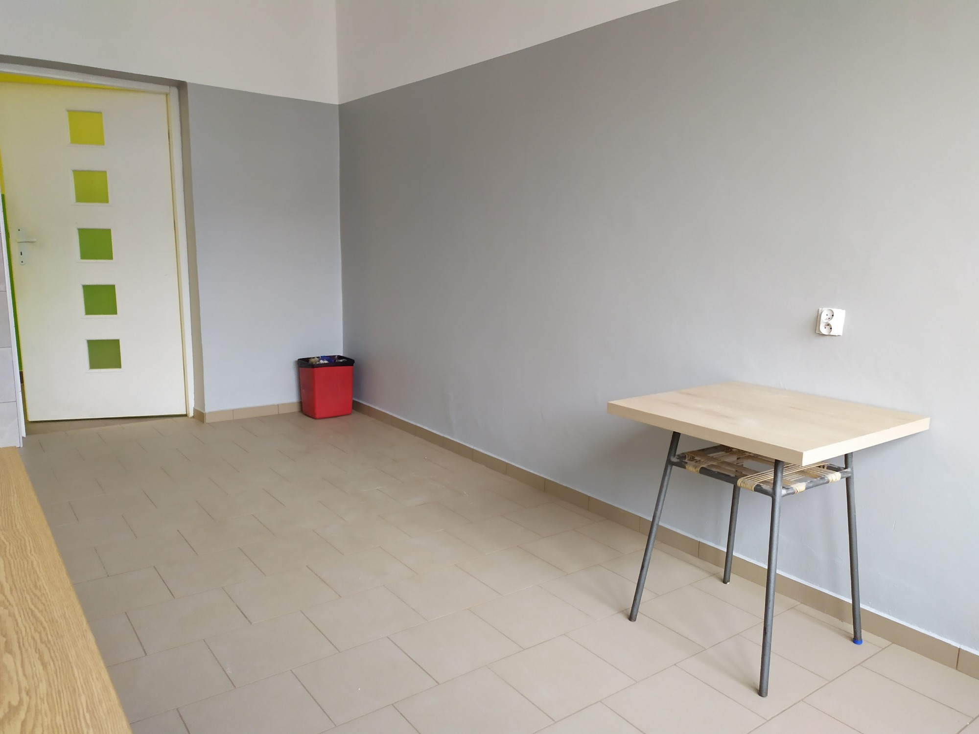 Szare ściany w pokoju po świeżym pomalowaniu, widoczna biała listwa przypodłogowa, brązowe płytki na podłodze i biurko na metalowych nogach z drewnianym blatem.