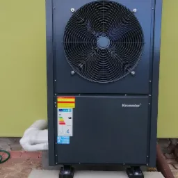 Pompa ciepła Krommler zamontowana przy ścianie budynku, z widoczną etykietą energetyczną i ocieplonym przewodem, na tle zielonej ściany.