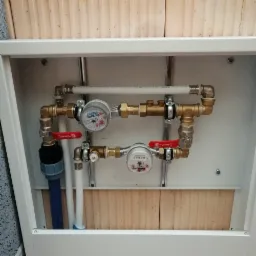 Dwa liczniki wody Zenner i Invena zamontowane w białej szafce z zaworami Perfect Water na tle drewnianej ściany.