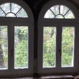 Dwa białe okna łukowe z widokiem na zieleń drzew, osadzone w surowej, ceglanej wnęce podczas remontu.