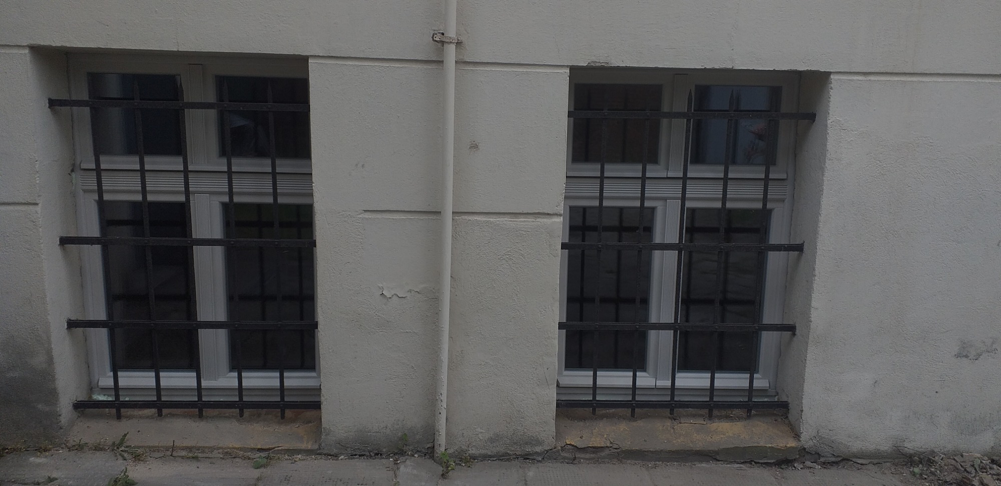 Dwa okna z białymi ramami zabezpieczone czarnymi, metalowymi kratami na tle szarej, teksturowanej ściany budynku z widocznym pionowym odpływem.