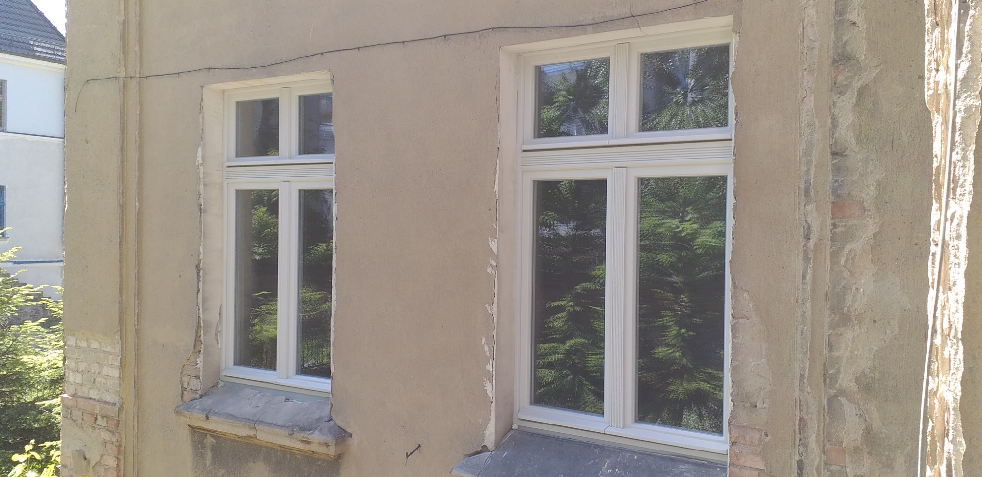 Dwa białe okna PCV w starym, częściowo odnowionym budynku z widocznymi uszkodzeniami tynku i cegłami, z odbiciem zieleni drzew w szybach.