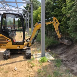 Żółta mini koparka CAT 301.7D podczas prac ziemnych, kopie rów w pobliżu metalowej konstrukcji.