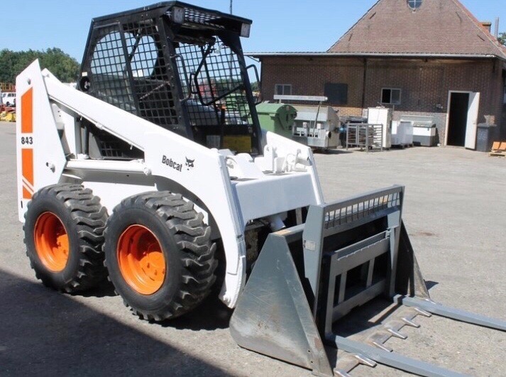 Biała ładowarka Bobcat 843 z pomarańczowymi felgami, widoczne widły i łyżka, na betonowym placu przed ceglanym budynkiem.
