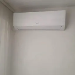 Biała jednostka klimatyzacyjna marki Gree zamontowana na białej ścianie, widoczna z lekkiego dołu, z zasłoną w lewym górnym rogu.