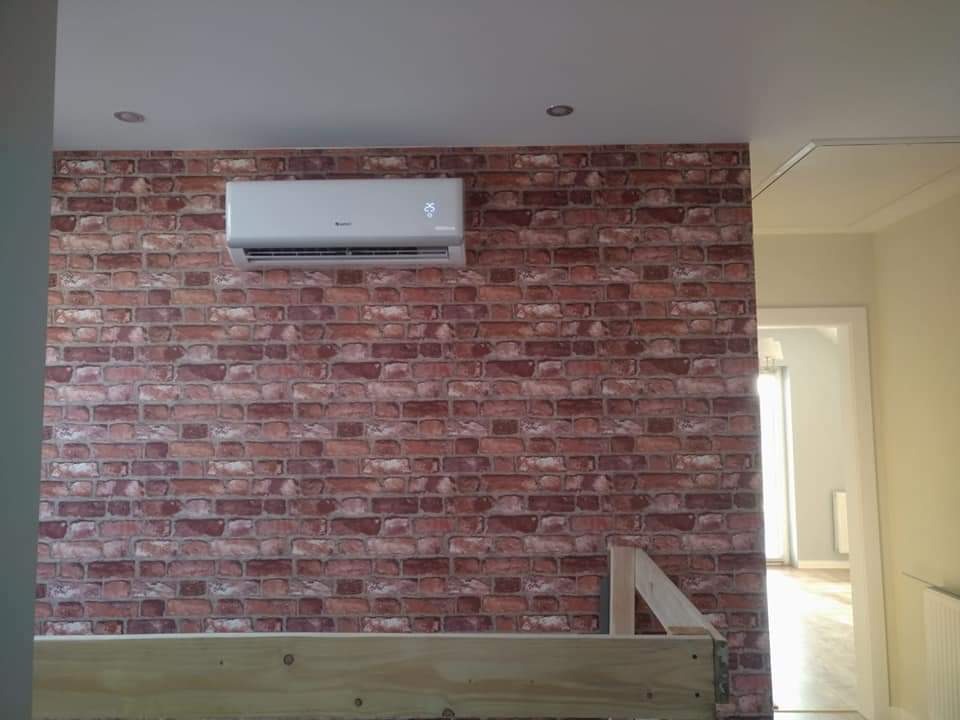 Szary klimatyzator zamontowany na ścianie z czerwonej cegły w pokoju z widoczną konstrukcją łóżka i otwartymi drzwiami do kolejnego pomieszczenia.