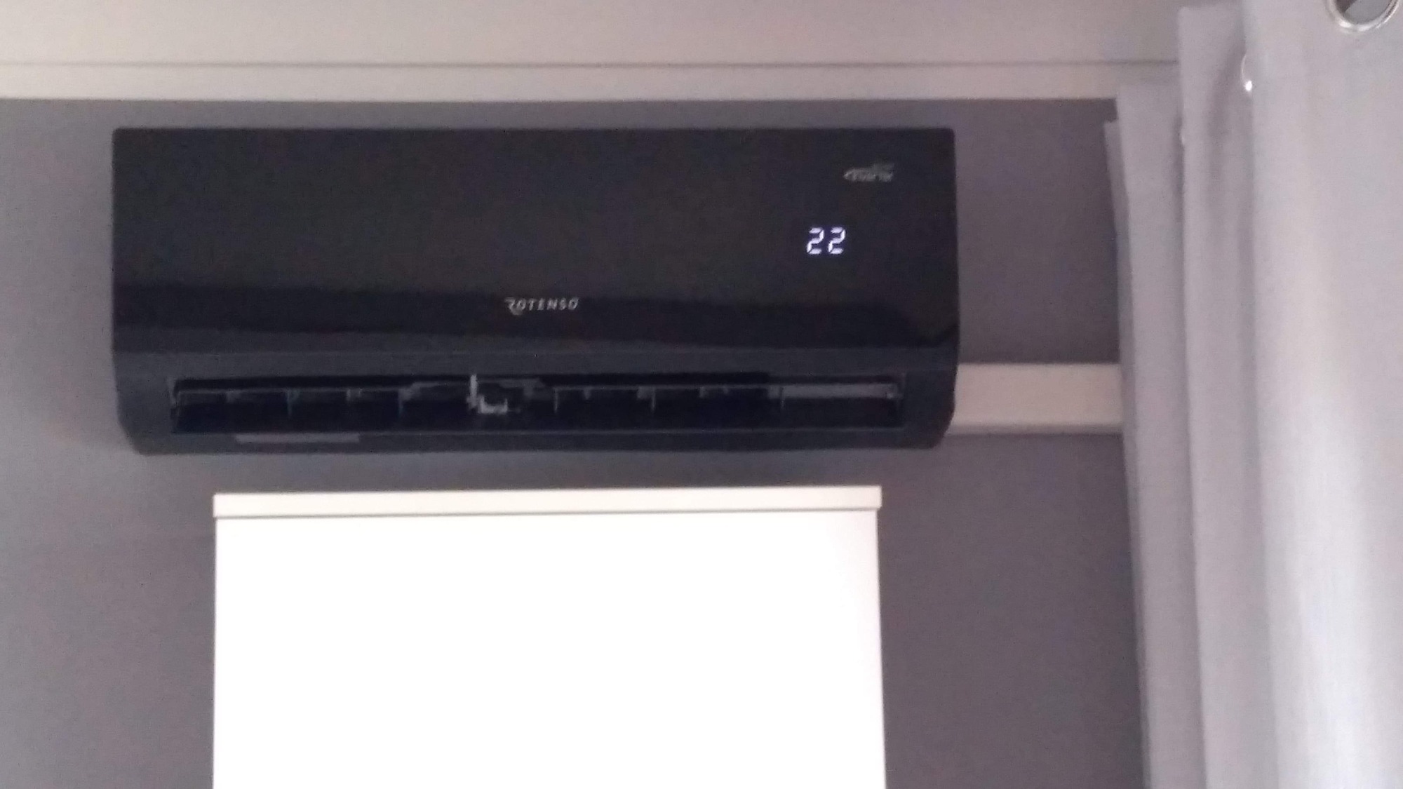 Czarny klimatyzator Rotenso zamontowany na szarej ścianie, wyświetlający temperaturę 22 stopnie, z fragmentem białej szafki i zasłony w tle.