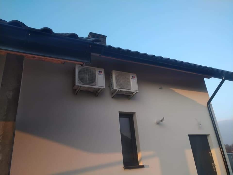 Dwa białe klimatyzatory zamontowane na ścianie budynku z ciemnym dachem i rynnami, widoczne okno i drzwi.