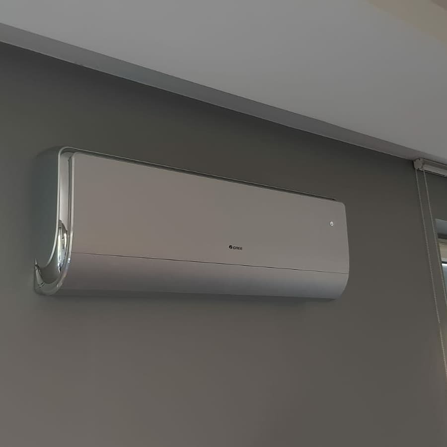 Nowoczesny, biały klimatyzator Gree zamontowany na szarej ścianie w pomieszczeniu z roletami okiennymi, widok z dołu.