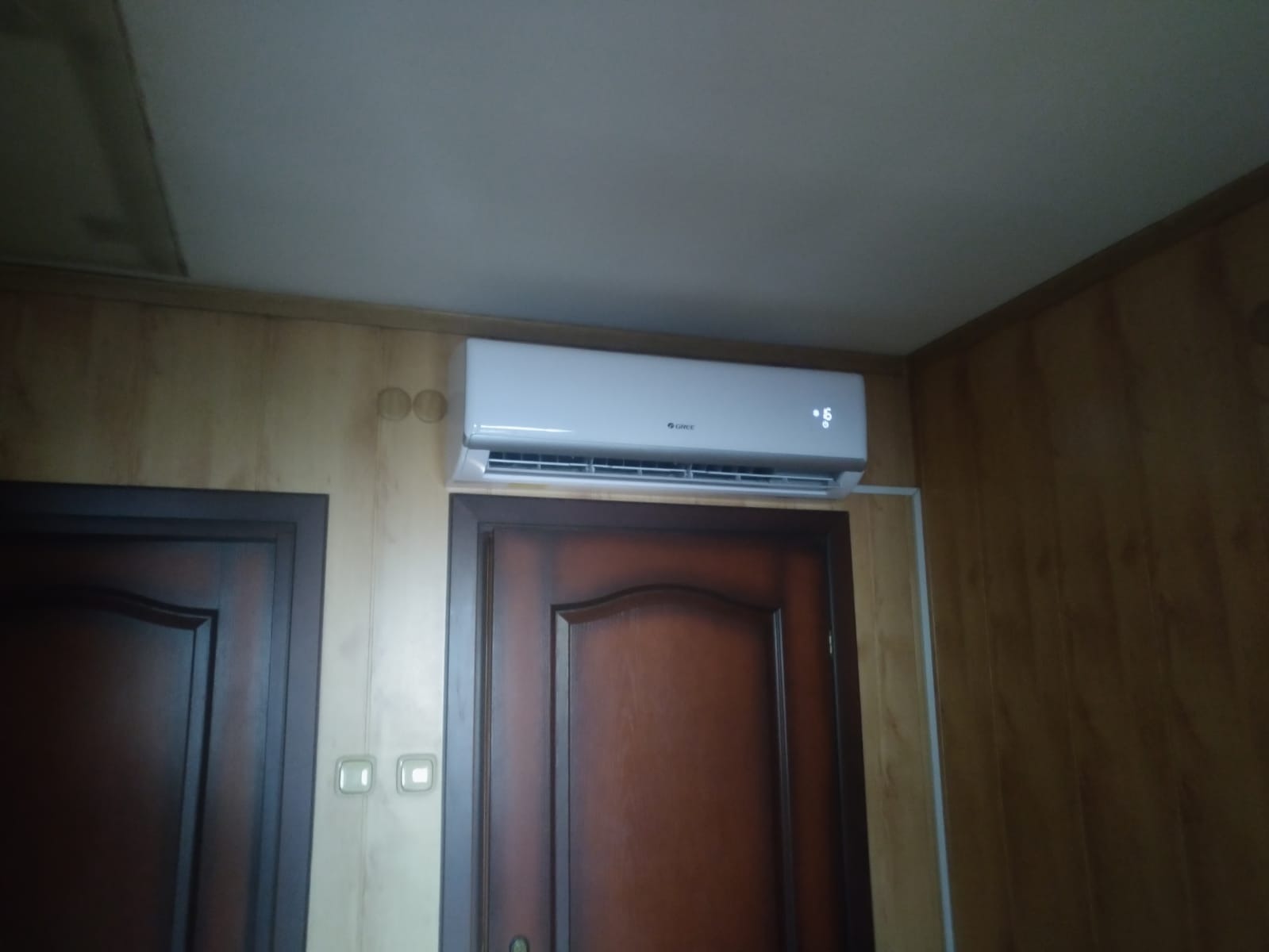 Biały klimatyzator Gree zamontowany nad ciemnymi drzwiami w pomieszczeniu z boazerią, widoczny wyświetlacz temperatury.