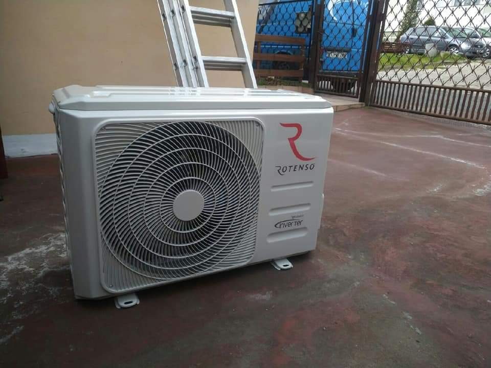 Biała jednostka zewnętrzna klimatyzacji Rotenso Inverter na brązowym podłożu, drabina aluminiowa i brama w tle.