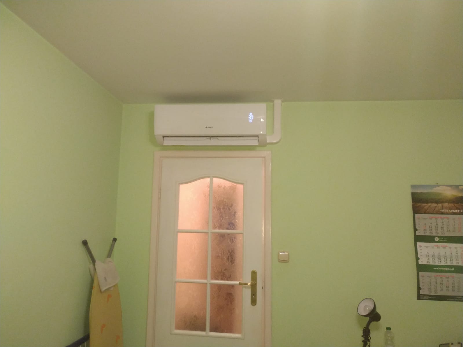 Biały klimatyzator zamontowany nad drzwiami w pokoju o jasnozielonych ścianach, widoczny kalendarz i deska do prasowania.