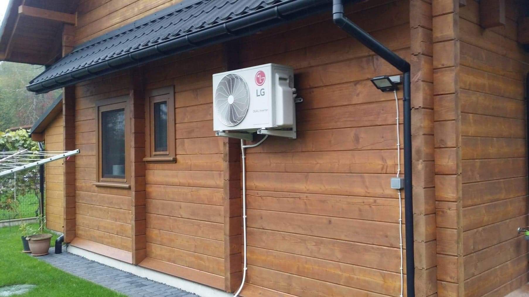 Zewnętrzna jednostka klimatyzacji LG zamontowana na drewnianej elewacji domu, z widocznymi przewodami i rurami odprowadzającymi skropliny.