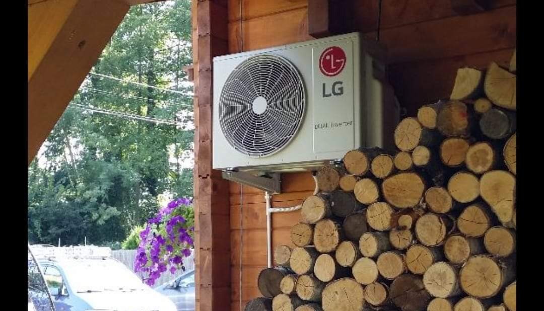 Zewnętrzna jednostka klimatyzatora LG zamontowana na drewnianej ścianie domu, obok stosu drewna opałowego, z widocznymi kwiatami i fragmentami samochodów w tle.