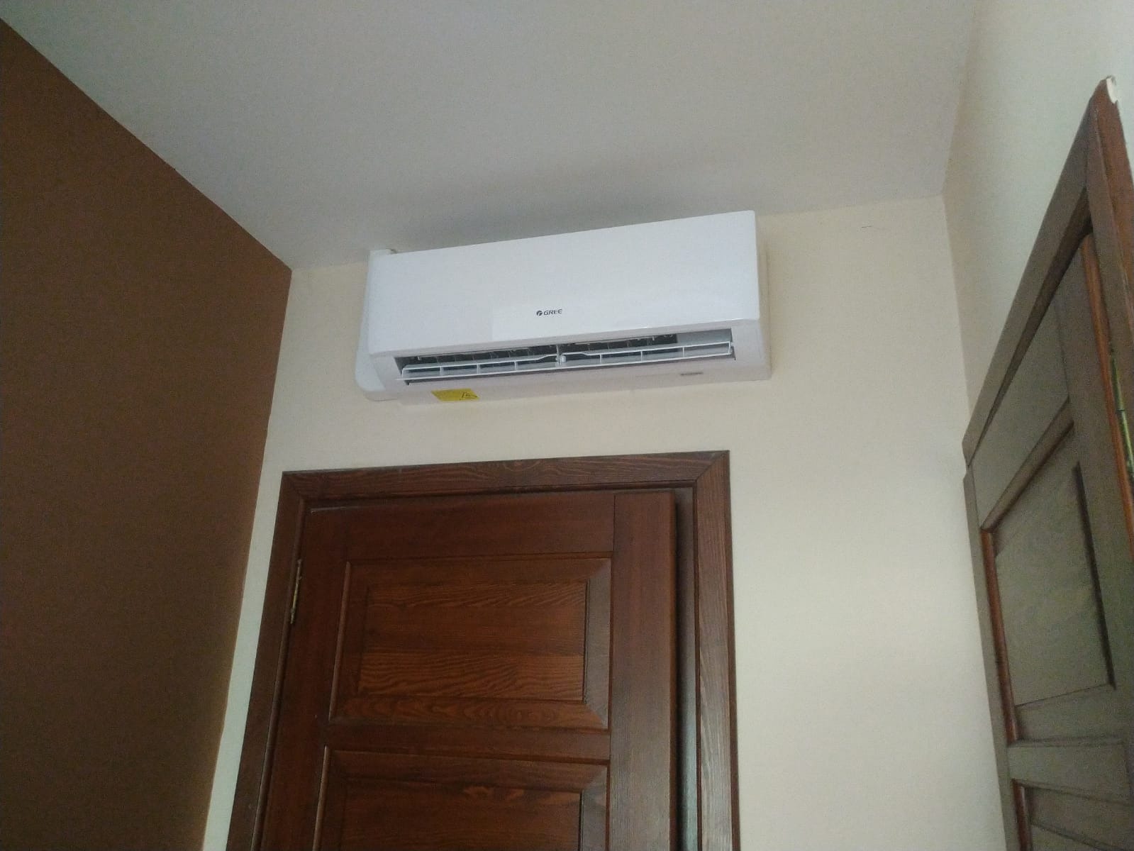 Biały klimatyzator marki Gree zamontowany nad mahoniowymi drzwiami w pokoju z brązową ścianą akcentującą.