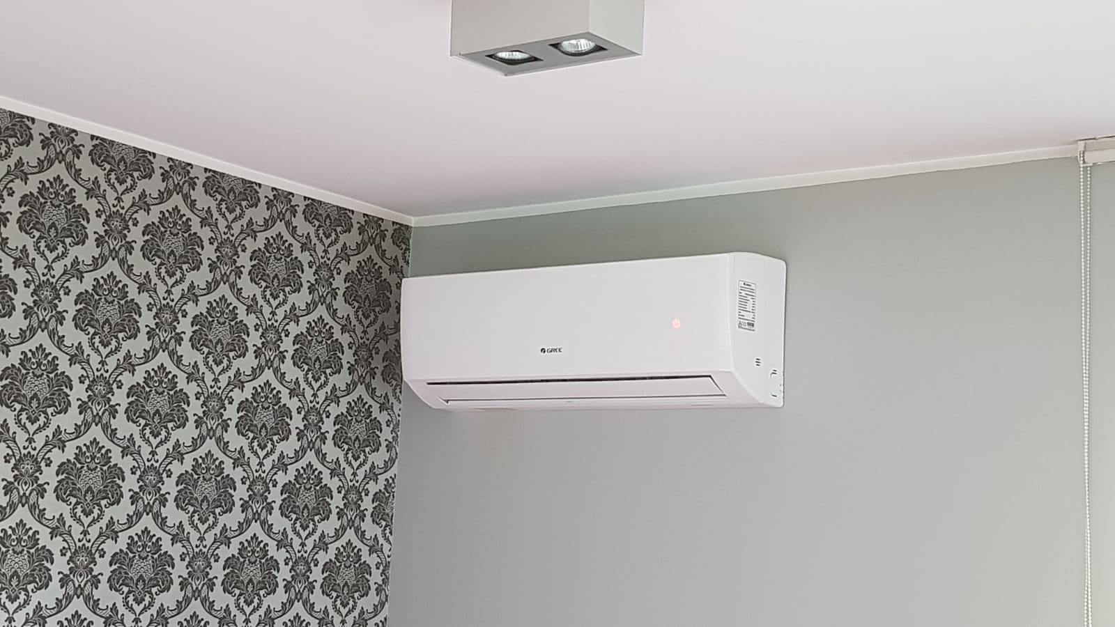Biały klimatyzator marki Gree zamontowany na szarej ścianie w pomieszczeniu z wzorzystą tapetą i nowoczesnym oświetleniem sufitowym.